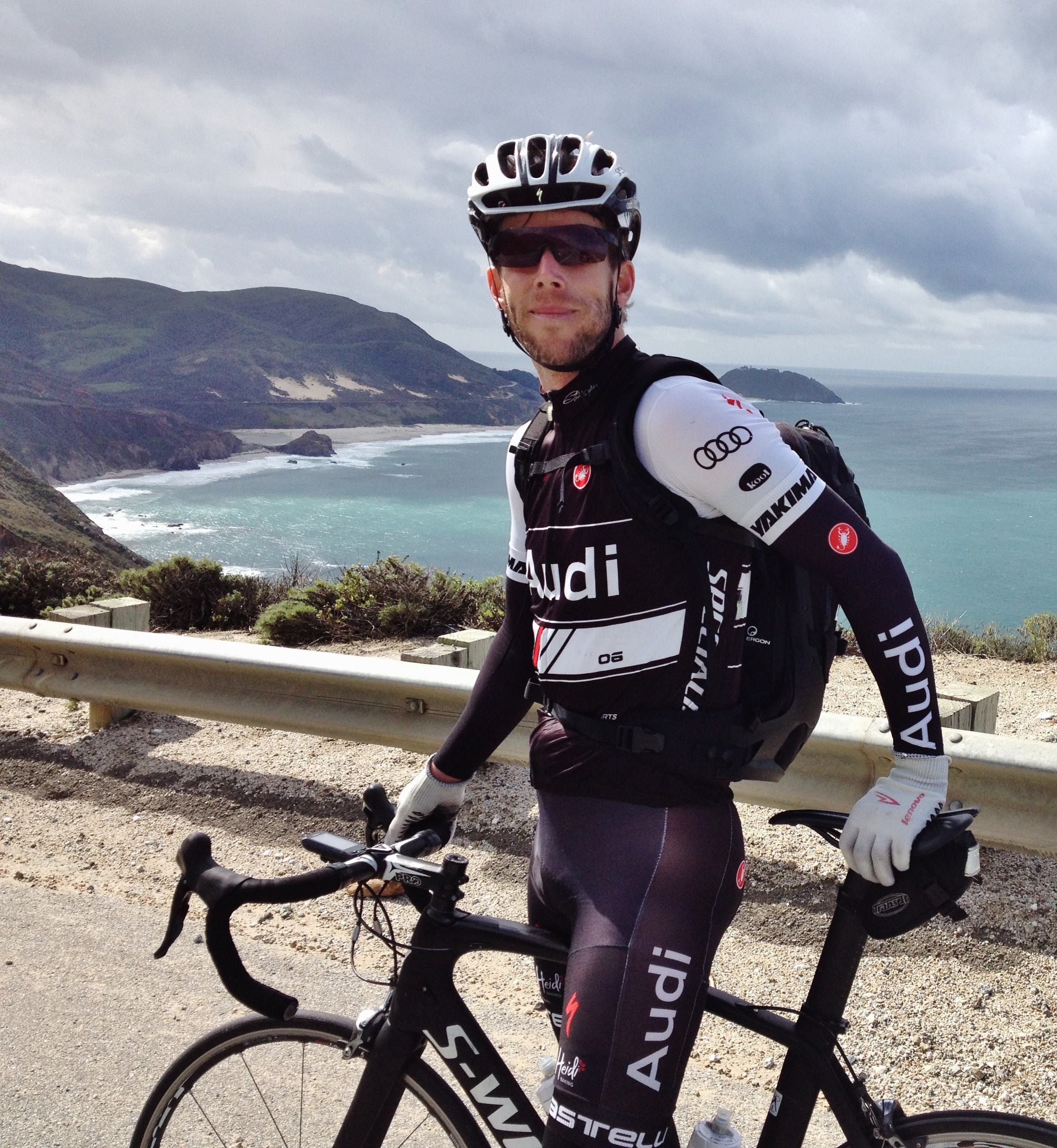 A cyclist in spandex stands with his bicycle above the Pacific ocean.